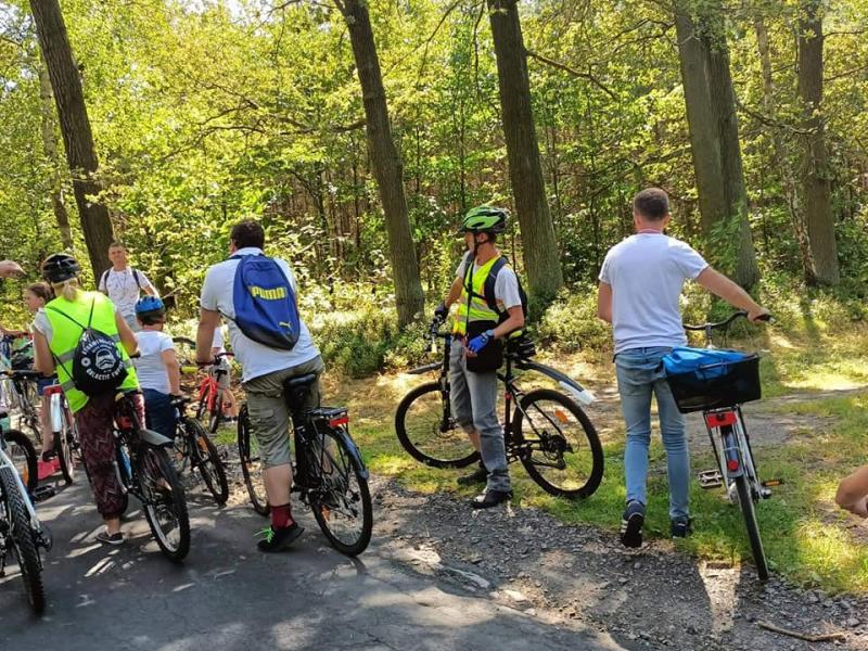 Na zdjęciu widzimy grupę rowerzystów, którzy przebywają w lesie. Grupa jest ubrana w stroje rowerowe i ma na twarzach wyraz radości i entuzjazmu. Widać, że wszyscy są gotowi do pokonania trasy rowerowej w otoczeniu natury. 