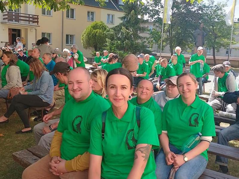 Na zdjęciu widzimy grupę ludzi i uczestników Środowiskowego Domu Samopomocy, ubranych w zielone koszulki, którzy siedzą na ławce wraz z innymi pielgrzymami. Siedzący uczestnicy są widoczni wzdłuż ławki, tworząc spójną grupę. Ich twarze wyrażają zainteresowanie, rozmowę i radość